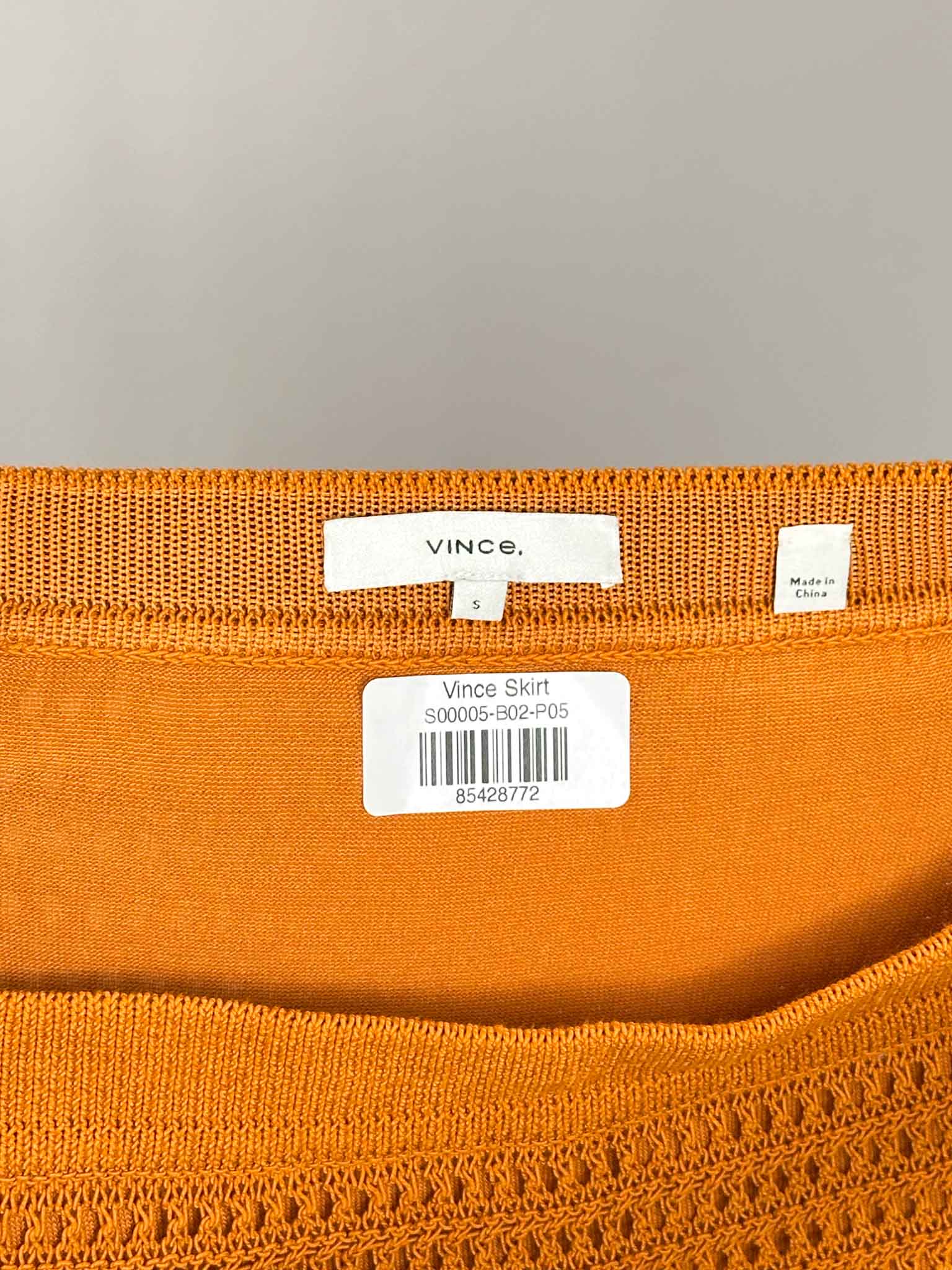 Close-up of an orange garment with a visible 'Vince' label on a gray background