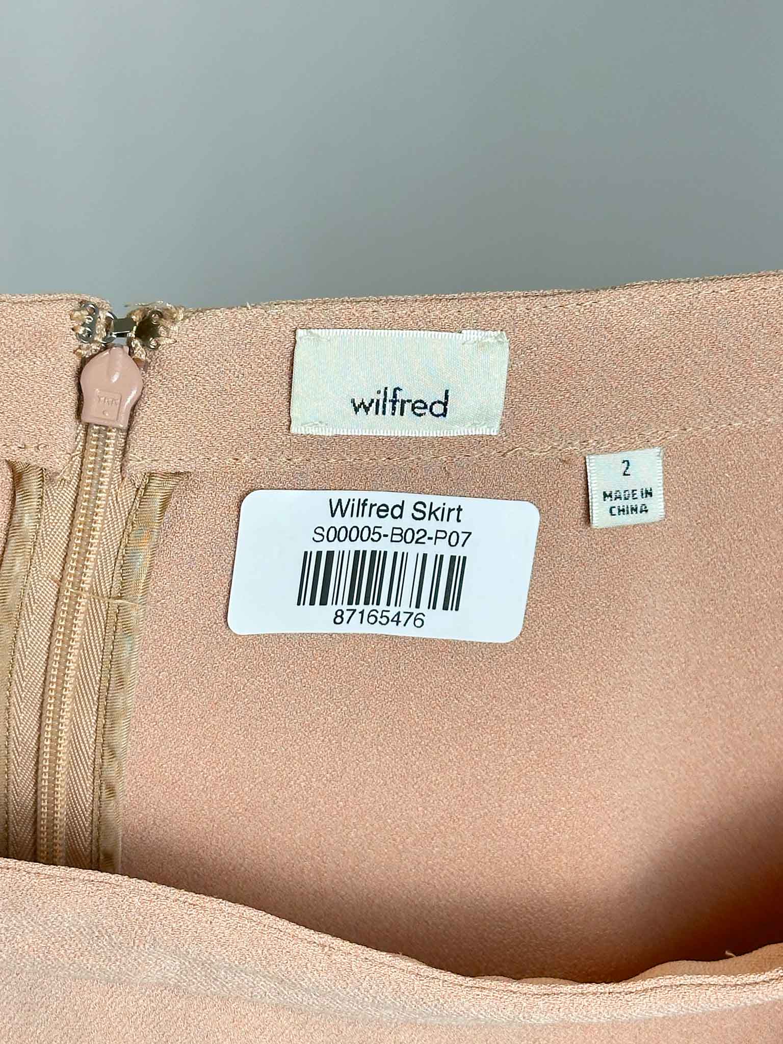 Close-up of a beige skirt with 'Wilfred' branding and labels on a gray background